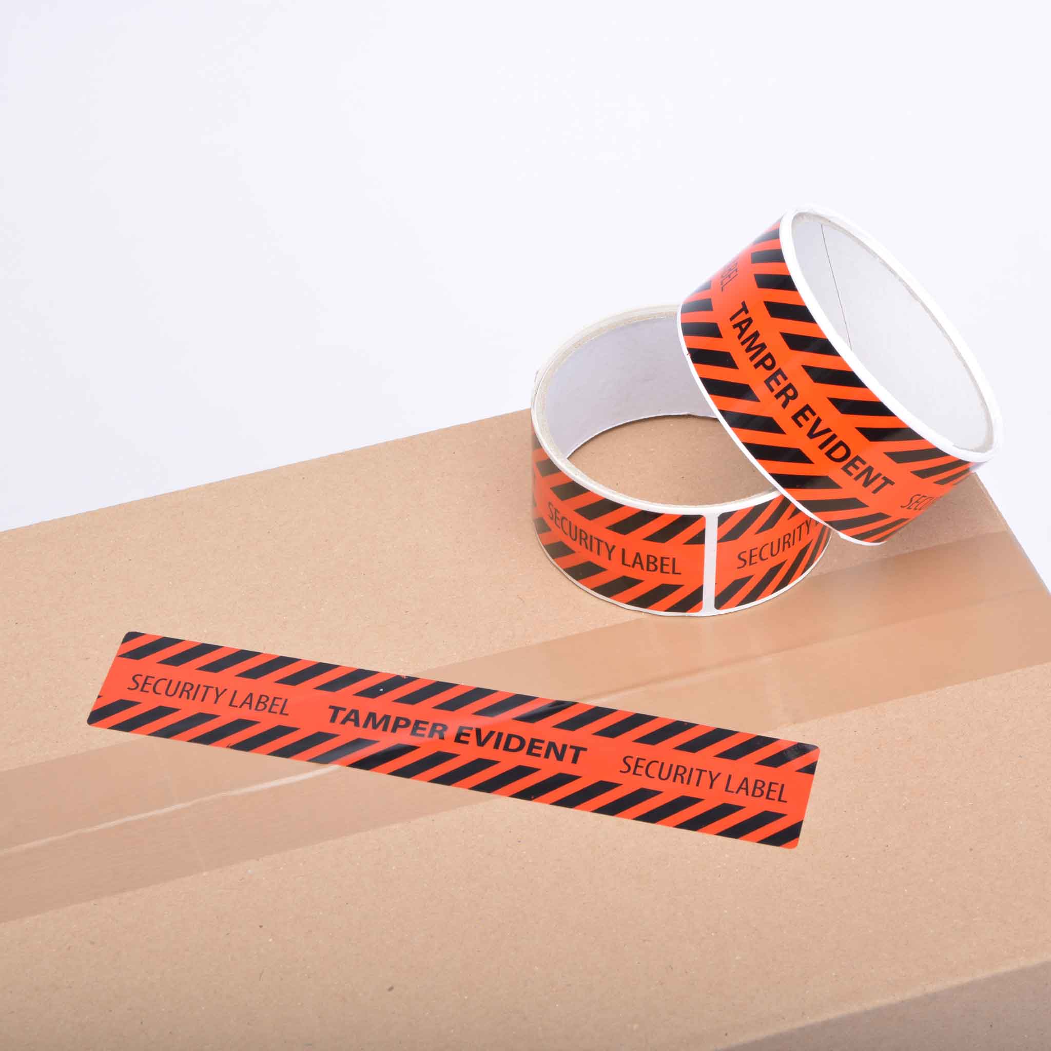 Sicherheitsetiketten Ein Karton von oben, auf dem zwei Rollen Sicherheitsklebeband liegen. Die Rollen sind schwarz/orange mit Schriftzug in der Mitte gestreift und haben einen weißen Kern. Ein Streifen ist auf dem Karton angebracht