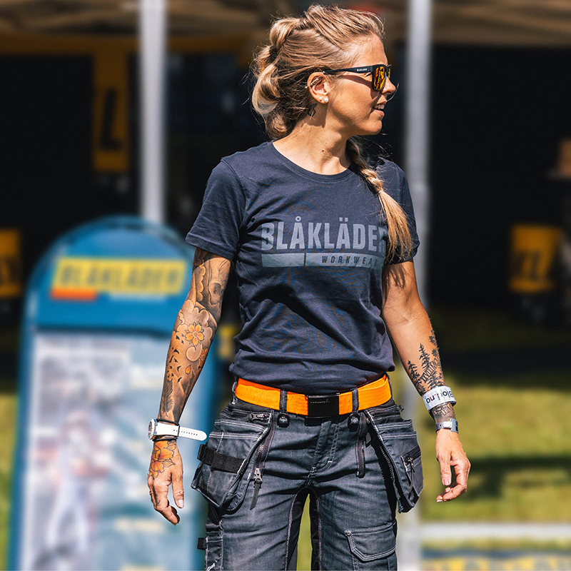 eine junge blonde Frau mit Sonnenbrille und einem dunkelblauen T-Shirt mit Blakläder Aufschrift in hellblau, einem orangenen Arbeitsgürtel und einer dunkelblauen Arbeitshose. Die Frau ist von vorn zu sehen und schaut grinsend auf die rechte Seite